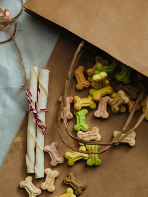 Aesthetic still life of colorful dog bone treats and rawhide chews spilling from a kraft paper bag, arrayed on a soft fabric.